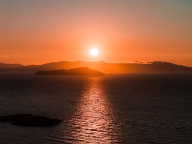 Yunanistan 'ın Girit Adası yakınlarındaki ufukta güneş batıyor. Gökyüzünde turuncu bir parıltı ve durgun bir su yansıtıyor. Uzakta görünen bir ada var..