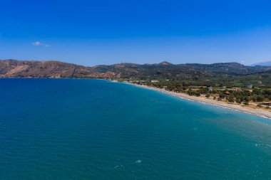 Yunanistan 'ın Girit Adası' nın havadan görünüşü turkuaz denizi, kumlu plaj, yemyeşil, dağınık binalar ve açık bir gökyüzü altında yuvarlanan tepeleri gösteriyor..
