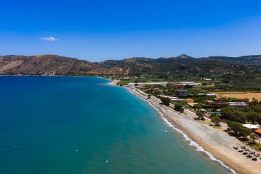 Yunanistan 'ın Girit Adası' nın havadan görünüşü, kumlu bir sahil, turkuaz sular, yeşillik, küçük binalar, tarım alanları ve kayan tepeleri gösteriyor.