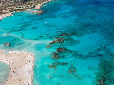 Yunanistan 'ın Girit kentindeki Elafonisi Sahili' nin hava manzarasında turkuaz sular, kumlu kıyı şeridi, güneşlenenler, şemsiyeler ve etrafındaki engebeli araziler görülüyor.