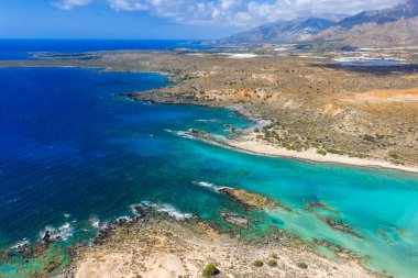 Yunanistan 'ın Girit kentindeki Elafonisi Sahili' nin hava manzarası, turkuaz ve mavi suları, sığ lagünleri, kayalık çıkıntıları ve bulutların altındaki uzak dağları gösteriyor..