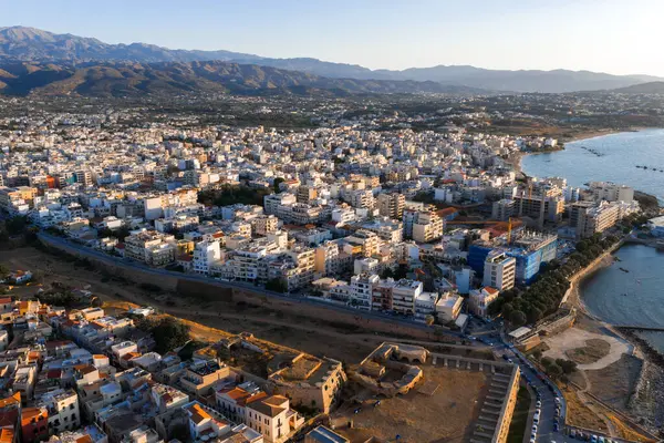 Günbatımında yukarıdan görülen Chania, Girit 'te beyaz ve pastel binalar, bir Venedik duvarı, berrak mavi sular ve uzakta engebeli dağlar bulunur..