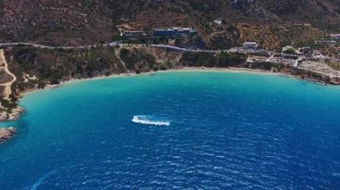 Girit 'teki Voulisma Sahili' nin havadan görünüşü, derin mavi sulara turkuvaz, engebeli tepeler, küçük bir yerleşim yeri ve beyaz izler bırakan tekneler gösteriyor..