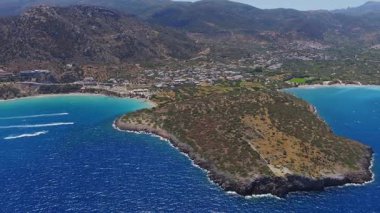 Girit 'teki Voulisma Sahili' nin havadan görünüşü turkuaz suları, engebeli uçurumları, hilal kıyı şeridini ve tekne veya jet ski hareketlerini gösteriyor..