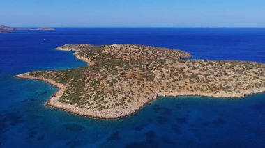 Girit 'in Agios Nikolaos kenti yakınlarındaki Akdeniz ile çevrili kayalık kayalıkları ve seyrek bitki örtüsü olan küçük bir adanın havadan görünüşü. Kamera pürüzsüzce hareket ediyor. Sahil şeridini ve açık gökyüzünün altındaki uzak tepeleri gösteriyor..