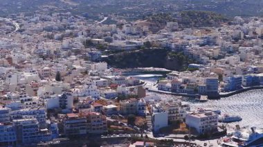 Girit, Agios Nikolaos 'un hava görüntüsü beyaz ve pastel binaları, yatları, mavi deniz suları ve çevresindeki tepeleri gösteren bir marina..