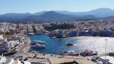 Girit 'teki Agios Nikolaos' un hava manzarası dairesel bir limanı, beyazlatılmış binaları, terasotta çatılarını, tekneleri, yeşillikleri ve engebeli dağları gösteriyor..