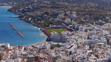 Girit, Agios Nikolaos 'un hava görüntüsü, mavi suları, yatlı bir marinayı, kumlu bir sahili, dalgakıranı ve körfezde uyanan tekneleri gösteriyor..