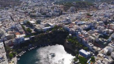 Beyaz ve pastel binalar, yemyeşil yemyeşil ve engebeli dağları berrak bir gökyüzü altında gösteren yavaş, süpürgen bir Agios Nikolaos, Girit tavası..