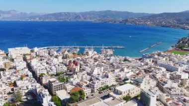 Girit 'teki Agios Nikolaos' un hava görüntüsü yat, iskele, pastel binalar, engebeli dağlar ve suda hareket eden bir tekne gösteriyor..