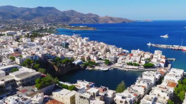 Girit, Agios Nikolaos 'un hava manzarası, Voulismeni Gölü, pastel binalar, bir marina, gezinti alanları, engebeli tepeler ve Akdeniz' i gösteriyor..