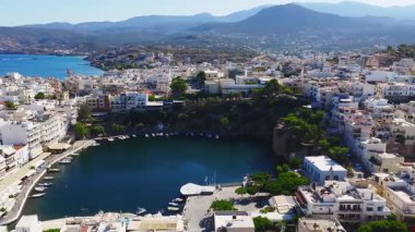 Girit 'teki Agios Nikolaos' un hava görüntüsü, beyazlatılmış binalar, dairesel bir göl, küçük tekneler, yuvarlanan tepeler ve Akdeniz kıyı şeridini gösteriyor..