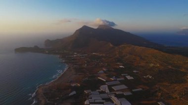 Girit 'in engebeli dağlarının, teraslı tepelerin ve seralarla kaplı tarlaların havadan görünüşü, altın günbatımı ışığı denizle ve uzak adayla buluşurken....