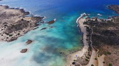 Elafonisi Sahili, Girit 'te yavaş bir hava temizliği, kıyı boyunca pembe kum, turkuaz sular, kayalık adalar, patikalar, ziyaretçiler ve şemsiyeleri gösteriyor..