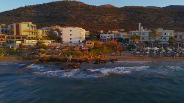 Stalida, Girit 'in üzerindeki havalimanında beyazlatılmış binalar, bir havuz, otopark ve gün batımının sıcacık renklerinde parlayan engebeli bir yamaç görülüyor..