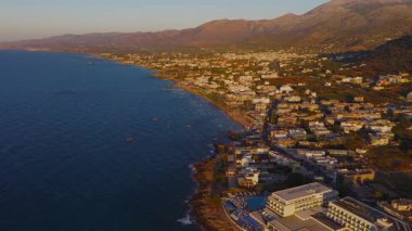 Günbatımında Stalida, Girit 'in kumlu sahillerini, tekneleri, beyaz binaları ve altın ışıklı dağları gösteren geniş bir hava manzarası..