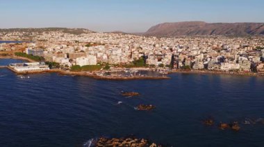 Yunanistan 'ın Girit kentindeki Chania kasabasının havadan görünüşü, limanı, rıhtımı, tekneleri, kentsel alanı, yeşilliği ve altın güneş ışığı altındaki dağları gösteriyor.