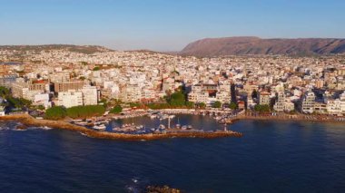 Girit, Chania 'nın havadan görünüşü, beyaz ve pastel binalar, bir marina, mavi deniz ve yavaş hareket eden engebeli dağları gösteriyor..