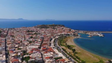 Tekneler, feribotlar, kumlu plajlar, palmiye ağaçları ve hareketli gemilerle dolu Rethymno limanının havadan görüntüsü..