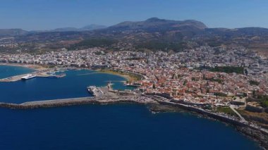 Girit, Rethymno 'nun havadan görünüşü, beyaz ve terakotta çatılı binaları, tekneli bir limanı, deniz fenerini ve arka planda engebeli dağları gösteriyor..