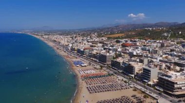 Girit, Rethymno 'nun havadan görünüşü, şemsiyeli kumlu bir sahil, hareketli bir gezinti alanı, tekneli turkuaz sular ve berrak bir gökyüzünün altındaki tepeleri gösteriyor..