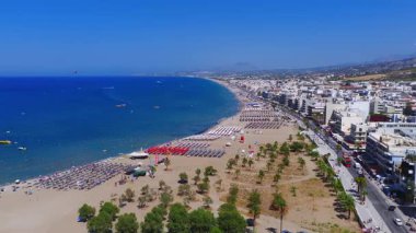Girit, Rethymno 'nun havadan görünüşü, şemsiyeli kumlu bir sahil, yüzücüler ve botlarla turkuaz suları ve arka plandaki kasabayı gösteriyor..