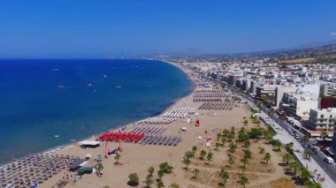 Rethymno 'nun kumlu sahilinin havadan görünüşü, şemsiyeler, yüzücüler ve paraşütçüler. Arka planda modern binalar ve tepeler olan gezinti alanı.