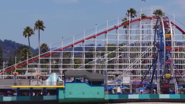 Kaliforniya 'daki Santa Cruz Sahili Boardwalk manzarası, Dev Ayı Lunaparkı hız treni, palmiye ağaçları, renkli binalar ve kıyı tepeleri yer alıyor..