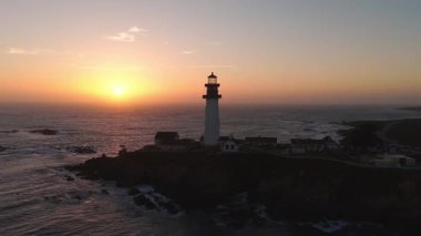 Güvercin Noktası Deniz feneri, günbatımında Kaliforniya 'da bir sahil uçurumunda duruyor. Dalgalar alttan vuruyor ve kamera dalgalı gökyüzünü yakalamak için dönüyor..