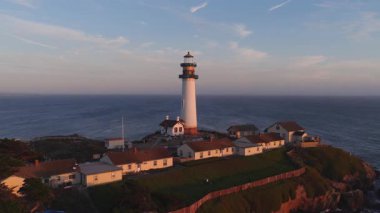 Güvercin Noktası Deniz feneri gün batımında engebeli bir uçurumun üzerinde durur. Etrafı altın ışıklı binalar, derin mavi okyanus ve yuvarlanan tepelerle çevrilidir. Küçük kamera hareketi.