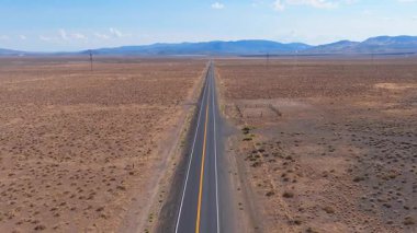 Nevada 'nın kurak çölündeki düz bir yolun hava manzarası, hareket halindeki araçlar, seyrek bitki örtüsü, elektrik hatları ve uzak dağ sıraları..
