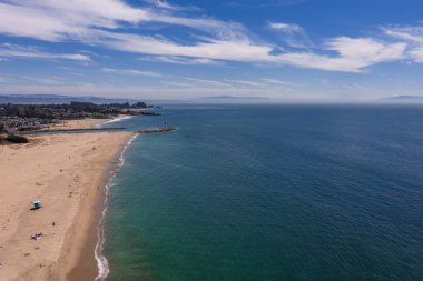 Santa Cruz, California 'nın havadan görünüşü, kumlu bir sahil, sakin Pasifik suları, bir iskele, dağınık ziyaretçiler ve bulutlu bir gökyüzünün altındaki uzak dağlar..