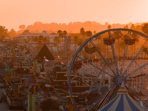 Santa Cruz Beach Boardwalk at sunset with the Giant Dipper rollercoaster, Ferris wheel, palm trees, and amusement park structures under an orange glow.