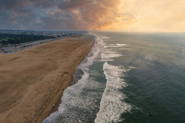 Expansive aerial view of a sandy beach with ocean waves, a glowing sunset sky, and a distant cityscape in San Francisco, California.