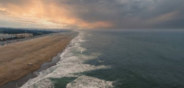 Kumlu kıyı şeridi, yumuşak dalgalar, binalar ve bulutları aşan altın güneş ışığı ile San Francisco, Kaliforniya 'daki Ocean Beach' in geniş hava manzarası.