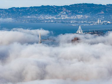 Aerial view of San Francisco partially covered in fog, featuring the Transamerica Pyramid, Bay Bridge, San Francisco Bay, and East Bay hills in the background.
