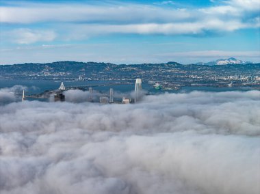Aerial view of San Francisco featuring the Bay Bridge above fog, Salesforce Tower, skyscrapers, and East Bay hills under a clear blue sky.