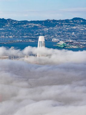 Aerial view of San Francisco featuring Salesforce Tower above fog, Bay Bridge, San Francisco Bay, port activity, rolling hills, and urban sprawl.