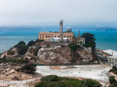 Alcatraz Island in San Francisco Bay features the historic prison and lighthouse atop rocky terrain, surrounded by blue waters and distant landforms.