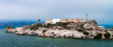 Alcatraz Island in San Francisco Bay features the historic penitentiary, a lighthouse, rugged cliffs, and turquoise waters under a cloudy sky.