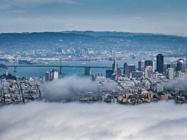 Aerial view of San Francisco featuring the Transamerica Pyramid, downtown skyscrapers, Bay Bridge, fog covered cityscape, and distant hills.