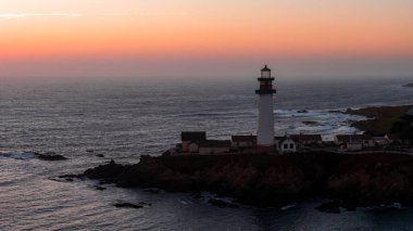 Aerial view of Pigeon Point Lighthouse on a rocky coastal outcrop in California, with ocean waves and a vibrant sunset sky in the background.