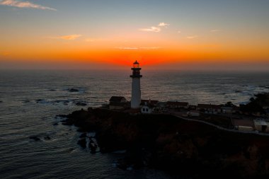 Pigeon Point Lighthouse stands on a rugged cliff in California, silhouetted by a vibrant sunset. The Pacific Ocean and rocky shoreline surround the scene.