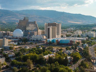 Reno, Nevada 'nın hava manzarası, şehir merkezindeki kumarhaneler, oteller, büyük küresel bir yapı, yerleşim alanları ve bulutlu bir gökyüzünün altındaki dağlar..
