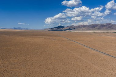 Expansive desert landscape with a straight road, sparse vegetation, and low mountains under a partly cloudy blue sky in Nevada.