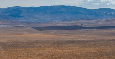 Expansive desert terrain in Nevada with dark rectangular industrial sections, sparse vegetation, and rugged mountains under a partly cloudy sky.
