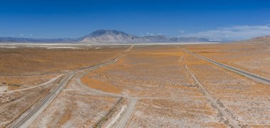 Expansive desert landscape with dirt roads, rail tracks, white salt flats, and a mountain range under a clear blue sky in Nevada.