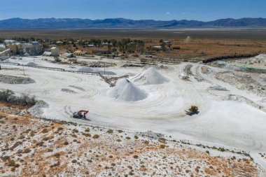 Aerial view of a Nevada salt mining site featuring white salt mounds, industrial machinery, small buildings, a train, and distant mountains.