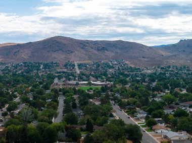 Aerial view of Carson City, Nevada, showing residential streets with greenery in the foreground and arid, rocky hills under a partly cloudy sky.