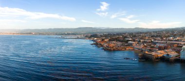 Aerial view of Monterey, California, featuring the coastline, Cannery Row piers, calm Pacific Ocean waters, rolling hills, and scattered clouds.